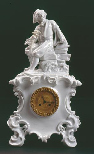 Γαλλικό επιτραπέζιο ρολόι με εκκερεμές από λευκή πορσελάνη του α μισού του 19ου αι. Διακοσμείται με τη μορφή Έλληνα νησιώτη καθισμένο σε βράχο θέμα επαναλαμβανόμενο σε χρηστικά αντικείμενα φιλελληνικής διακόσμησης.