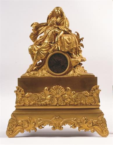 Επιτραπέζιο ρολόι με εκκρεμές, από επιχρυσωμένο ορείχαλκο. Διακοσμείται με την αλληγορική παράσταση του Λόρδου Μπάυρον στην αγκαλιά της Ελλάδος. Είναι έργο του οίκου C.F.Petit και χρονολογείται γύρω στο α μισό του 19ου αι.