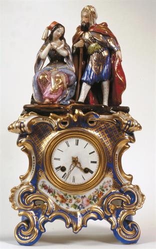 Επιτραπέζιο ρολόι με εκκρεμές από πολύχρωμη πορσελάνη. Διακοσμείται με ολόγλυφη  ρομαντική σύνθεση  μιας νεαρής ελληνίδας και ενός ανατολίτη.