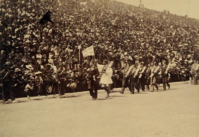 Ολυμπιακοί Αγώνες 1896. Παρέλαση Ολυμπιονικών στο Παναθηναϊκό Στάδιο. Προηγείται ο Σπύρος Λούς, νικητής του Μαραθωνίου δρόμου κρατώντας την ελληνική σημαία. Μπροστά του ο Κωνσταντίνος Μάνος. Φωτογραφία.
